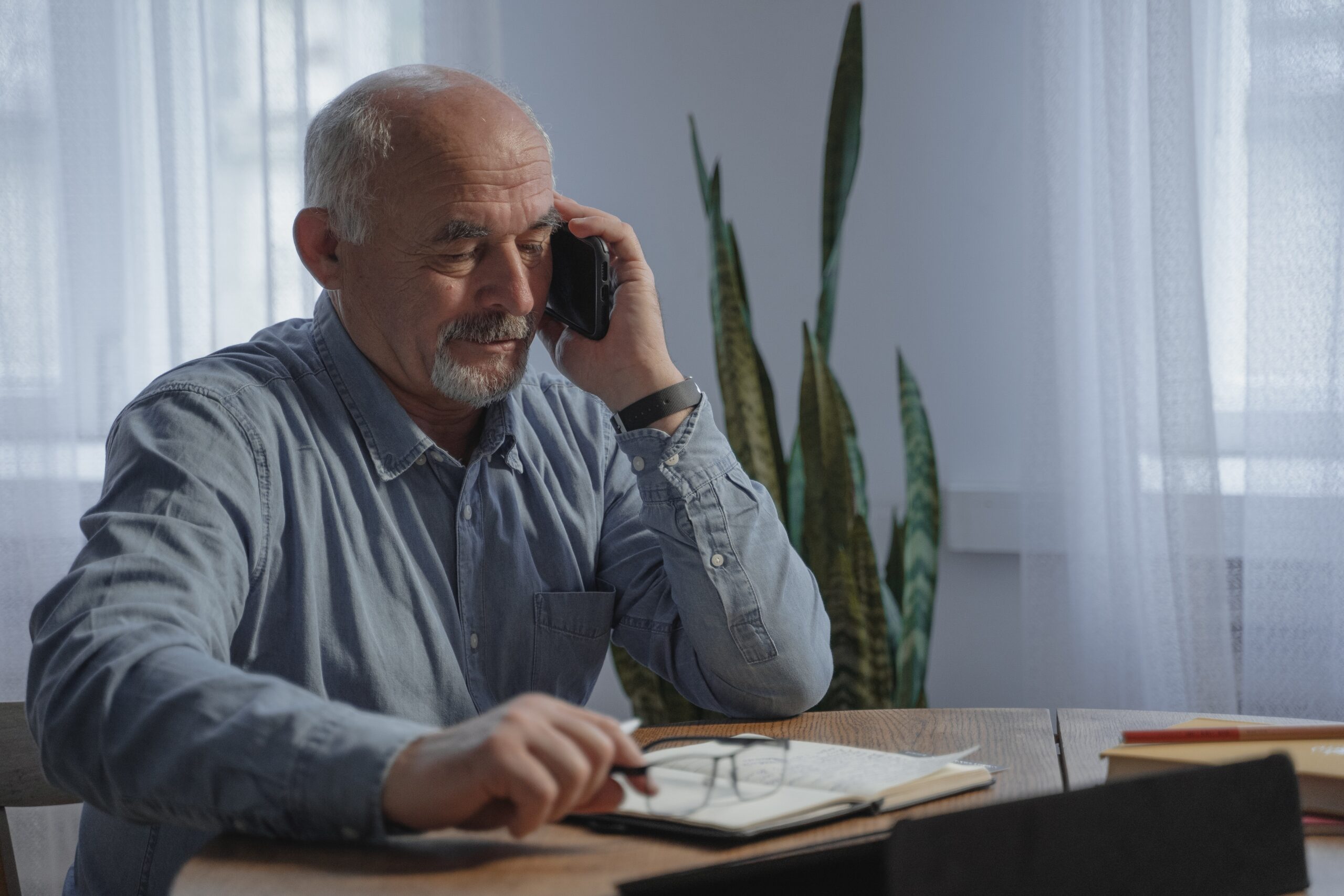 Older gentleman sat at a table with a note-pad on the table with pen and phone chatting.