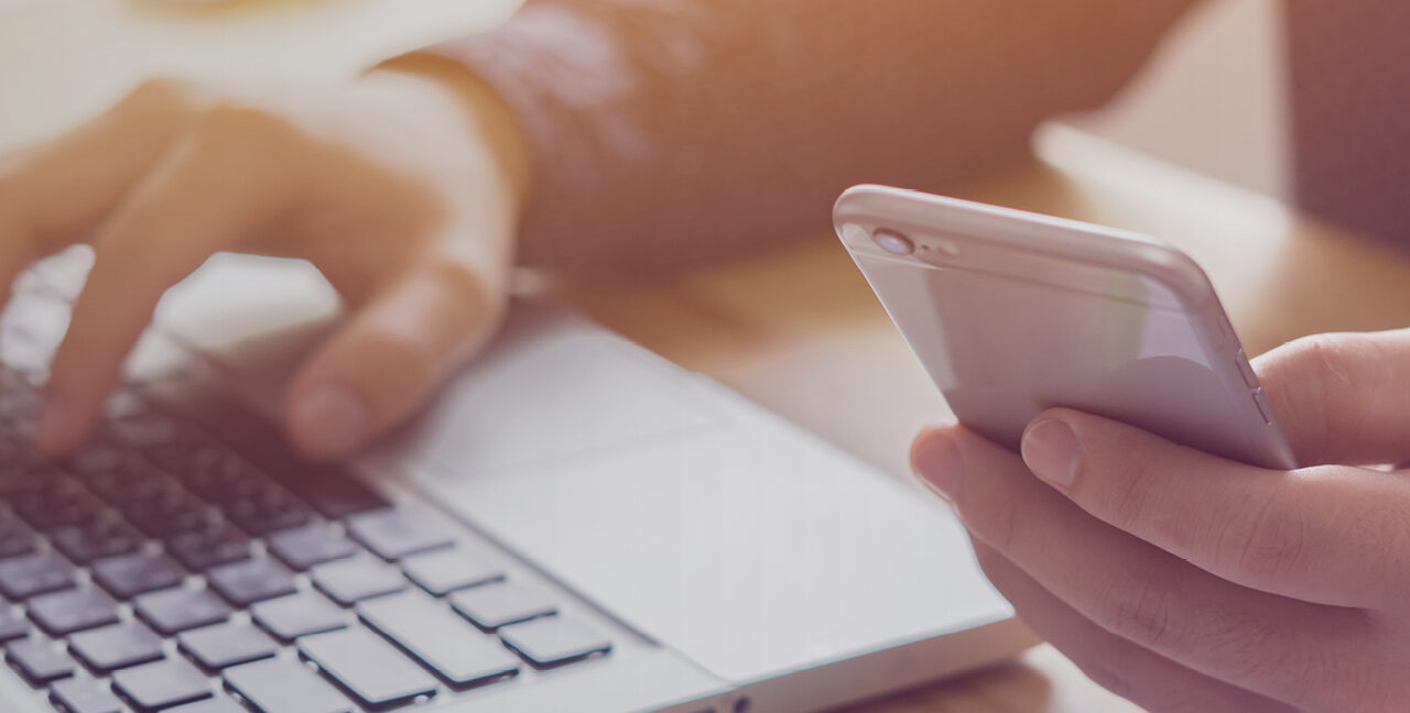 Person sat at a desk with one hand on a laptop and the other using a mobile phone.