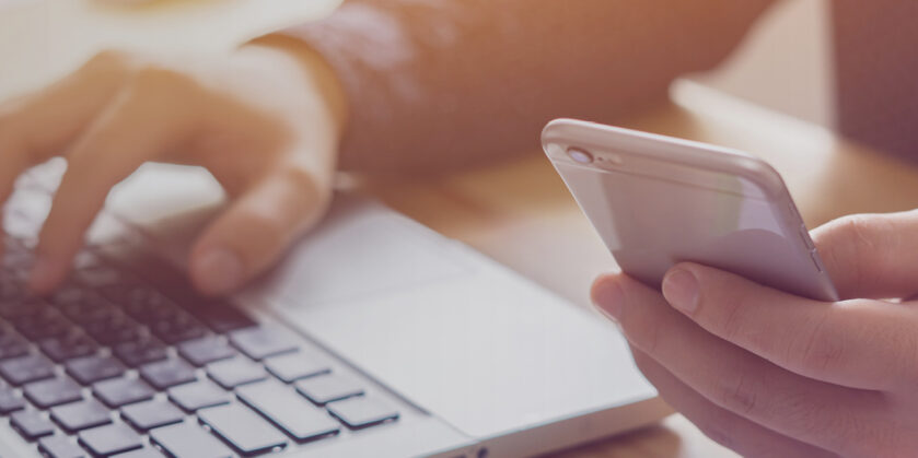 Person sat at a desk with one hand on a laptop and the other using a mobile phone.