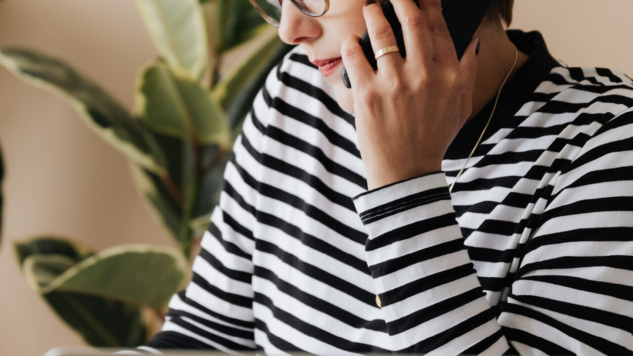 Woman holding a mobile phone up to her left ear. She is wearing a black and white stripy top and wears glasses.