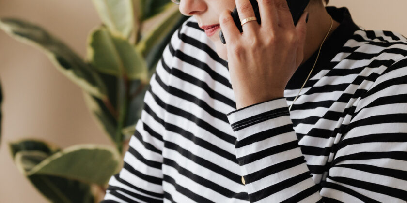 Woman holding a mobile phone up to her left ear. She is wearing a black and white stripy top and wears glasses.