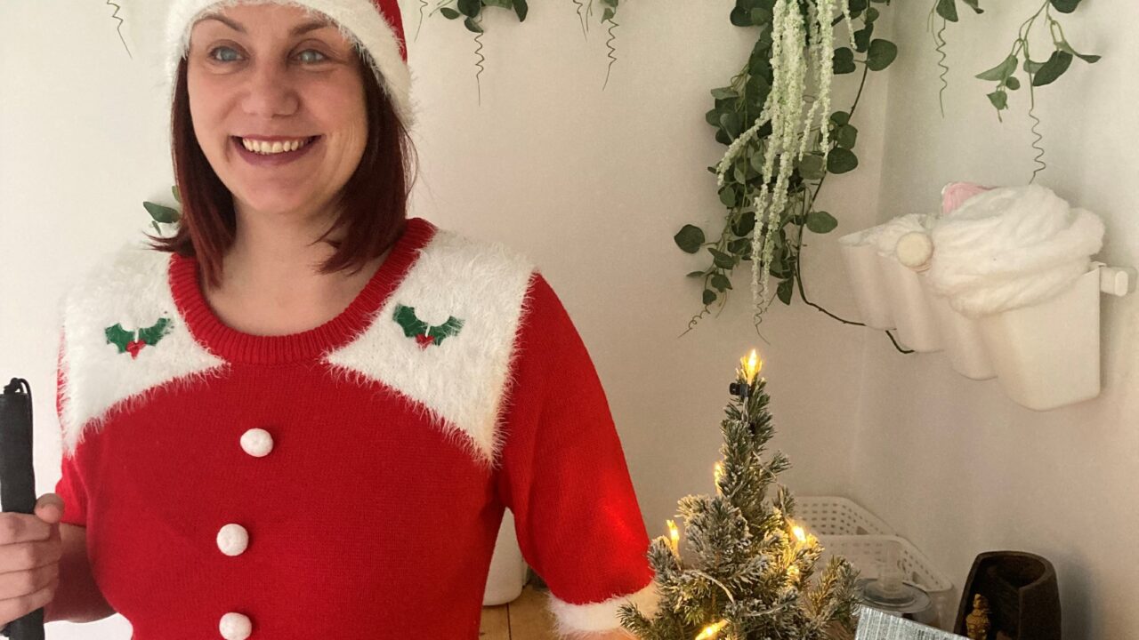 Nina is wearing a red festive hat and dress, standing in front of a table with a small lit Christmas tree, holding her cane