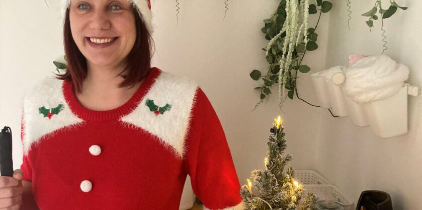 Nina is wearing a red festive hat and dress, standing in front of a table with a small lit Christmas tree, holding her cane