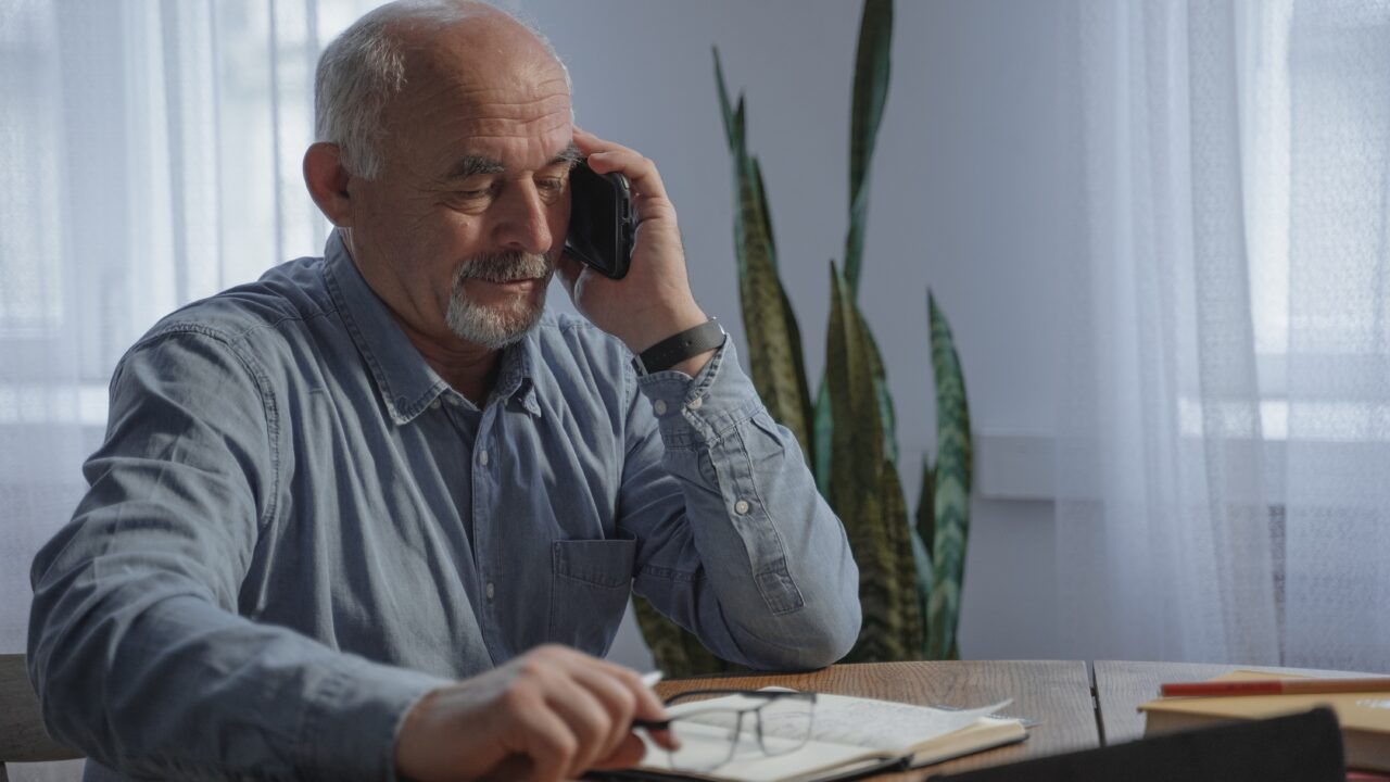 Older gentleman sat at a table with a note-pad on the table with pen and phone chatting.