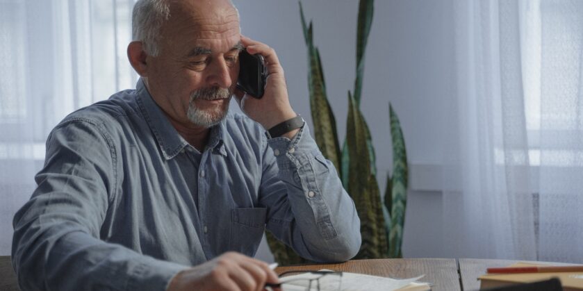 Older gentleman sat at a table with a note-pad on the table with pen and phone chatting.