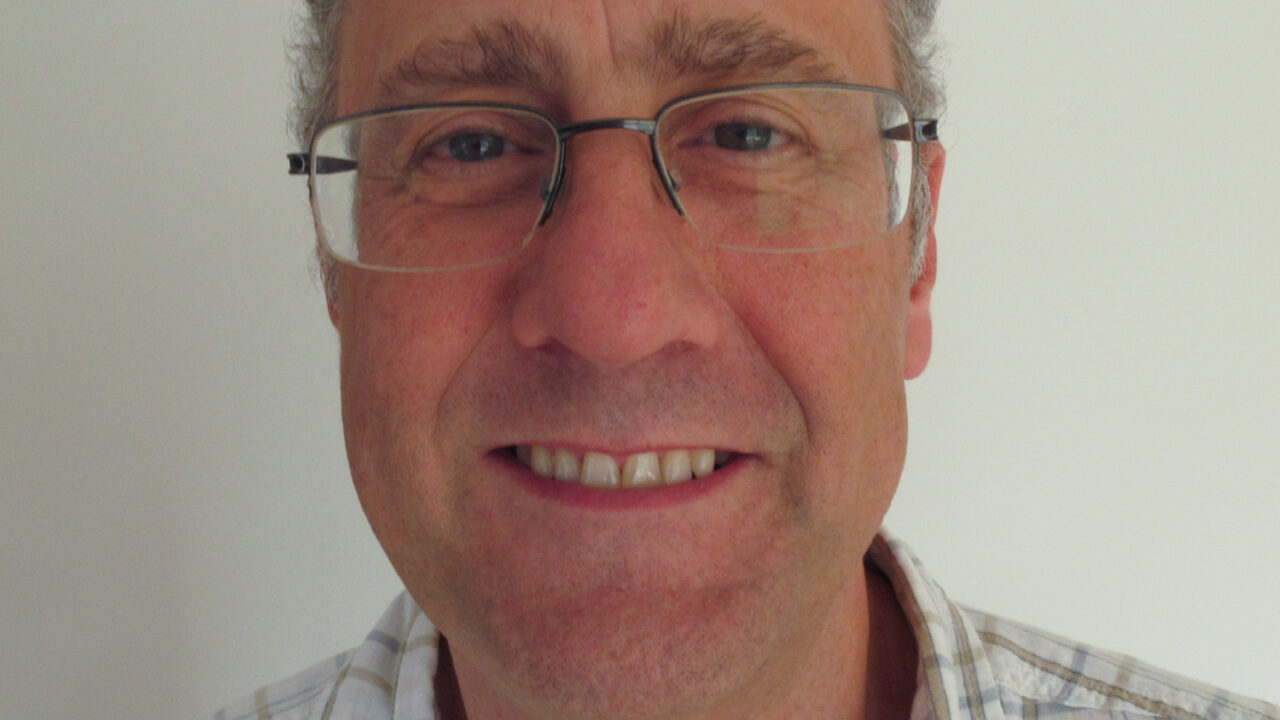 Image shows a picture of Carl Hodge. He is smiling towards camera and wears a checked shirt and glasses.
